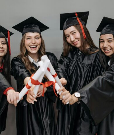 A joyful group of multiracial women in graduation gowns holding diplomas.